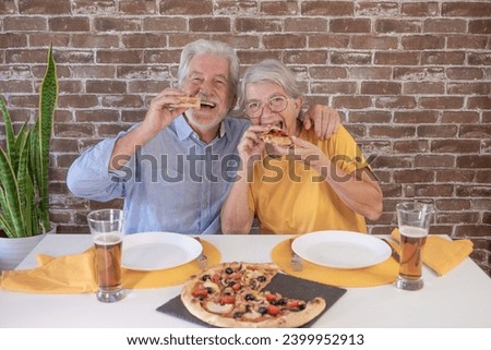 Happy senior couple looking at camera biting a slice of pizza. Elderly woman and man share a traditional Italian pizza for dinner