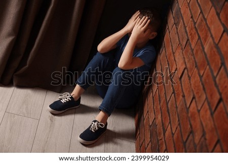 Child abuse. Upset boy sitting on floor near brick wall indoors Royalty-Free Stock Photo #2399954029