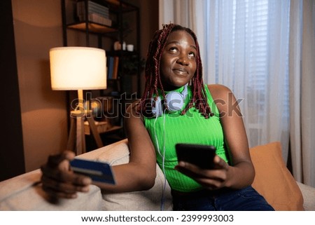 Curious delighted dreamy woman wearing casual colourful bright outfit dreaming looking up wearing new headphones holding cell phone and debir card in hands doing online purchase. Royalty-Free Stock Photo #2399993003
