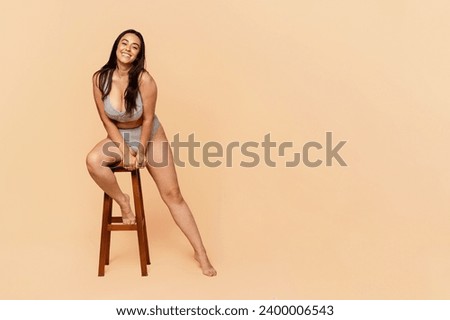 Young cheerful european lady with long hair, seated on wooden stool, laughs in relaxed pose, on light background, embodying joy and ease. Happiness, comfort, positive emotion