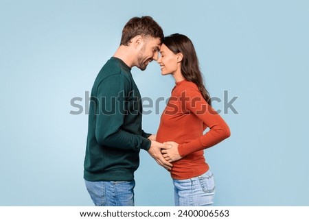 Affectionate and expectant young couple shares a close, tender moment as they both lovingly place their hands on the woman's pregnant belly against blue backdrop Royalty-Free Stock Photo #2400006563