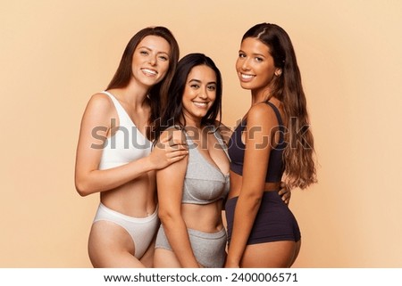 Three cheerful multiracial women in underwear embrace and smile against warm beige background, depicting camaraderie and diverse beauty. Friendship, diversity, joy and lifestyle Royalty-Free Stock Photo #2400006571