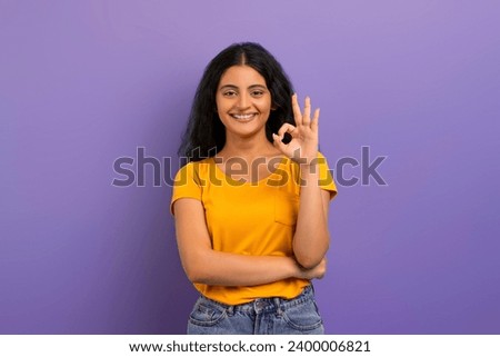 Positive cheerful pretty young indian woman wearing casual clothing showing okay gesture and smiling at camera, posing isolated on purple studio background. Everything just fine