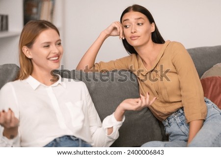 Envy In Friendship. Jealous Lady Listening To Her Cheerful Friend Talking And Bragging About Her Great Life Sitting On Couch At Home, Rolling Eyes During Conversation. Selective Focus Royalty-Free Stock Photo #2400006843