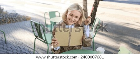 Portrait of young beautiful blond woman, artsy girl in coffee shop, holding notebook and pen, writing in her journal, drawing sketches outdoors.