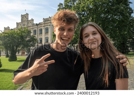 Cheerful young woman and man take photo together against campus. Young lady smiles while guy shows peace sign making funny faces