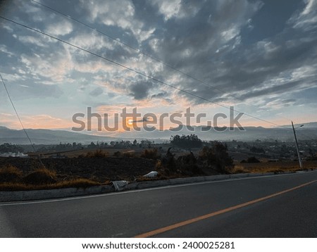 A SUNSET WHILE I WAS WALKING ON THE PUBLIC ROAD AND THE MOUNTAINS OF THE SIERRA AND AN ORANGE SUN WITH A PUBLIC LIGHTING AND PLANTS AROUND THE TREES WITH A BLUE SKY ALSO A CLOUDY GRAY SKY