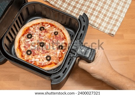 HAND BAKING A PIZZA IN AIR FRYER IN THE KITCHEN. ULTRA PROCESSED AND FAST FOOD CONCEPT. TOP VIEW.