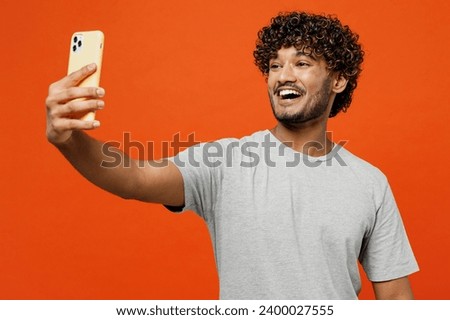 Young smiling happy Indian man he wears t-shirt casual clothes doing selfie shot on mobile cell phone post photo on social network isolated on orange red background studio portrait. Lifestyle concept
