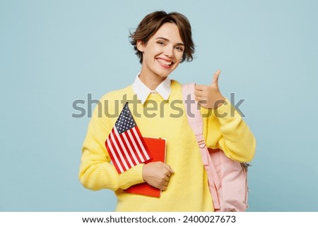 Young smiling happy woman student wear casual clothes yellow sweater backpack bag hold notebook American flag show thumb up isolated on plain blue background. High school university college concept