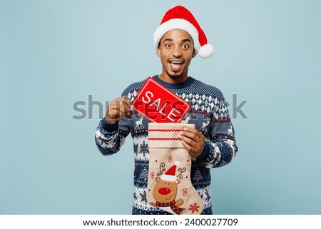 Young fun man wear sweater Santa hat posing hold stocking for gift, card sign with sale title text isolated on plain pastel blue background. Happy New Year 2024 celebration Christmas holiday concept