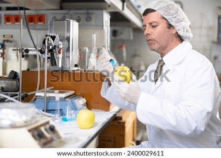 Biochemist performing GMO experiments in modern laboratory, injecting chemical substances into apple. High quality photo Royalty-Free Stock Photo #2400029611