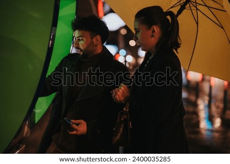 A couple uses an ATM on a rainy day. The man withdraws money while holding an umbrella. Business people are seen having a conversation. Royalty-Free Stock Photo #2400035285