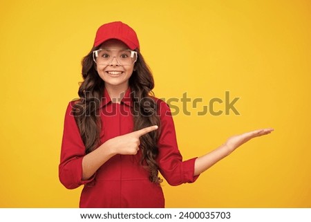 Worker teenager child wearing overalls red, cap and protect glasses. Studio shot portrait isolated on yellow background. Pointing and showing concept.