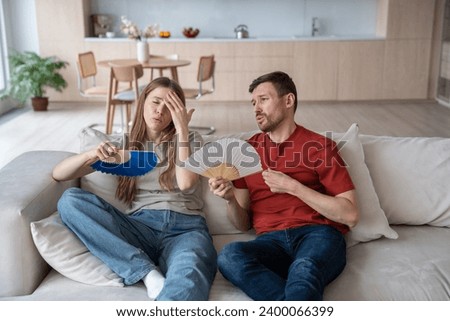 Overheated couple suffering from hot sultry air, waving with hand fans. Sweaty wife touching forehead, having headache, dehydrated husband with difficult breath. Necessity for conditioner in summer Royalty-Free Stock Photo #2400066399