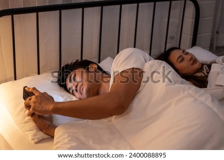 Asian man secretly chatting to mistress while lying in bed with girlfriend. Attractive disloyal husband using phone communicate with paramour lover while his wife sleeping late at night in bedroom. Royalty-Free Stock Photo #2400088895