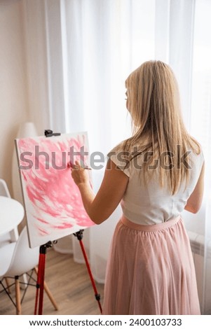 Pretty young blonde woman artist with palette and brush painting abstract pink picture on canvas at home. Art and creativity concept. High quality photo