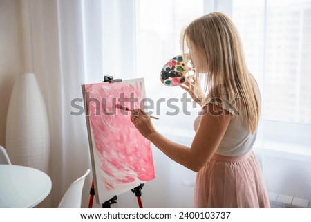 Young blonde woman artist with palette and brush painting abstract pink picture on canvas near window. Art and creativity concept. High quality photo