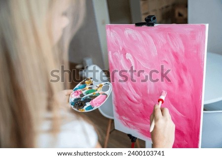 Back view of young woman artist with palette and brush painting abstract pink picture on canvas. Art and creativity concept. High quality photo