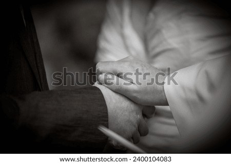 This emotive black and white photograph captures a close-up of two people holding hands in a comforting clasp. The hands, one clad in a sleeve of a white shirt and the other in a darker fabric, likely Royalty-Free Stock Photo #2400104083