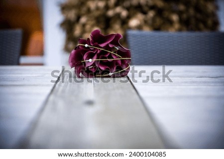 This evocative image captures a single purple flower, its petals slightly wilted, lying in the center of a rustic wooden table. The flower's deep hue stands out against the neutral tones of the wood Royalty-Free Stock Photo #2400104085
