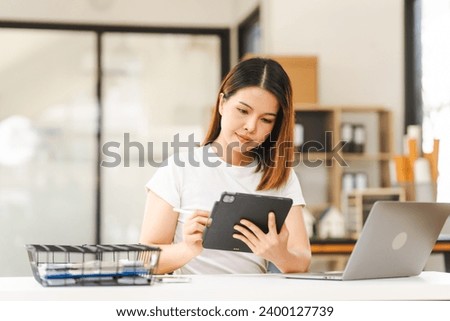 Cheerful Asian woman confidently uses mobile phone, tablet with laptop and notebooks on the desk in a bright office.