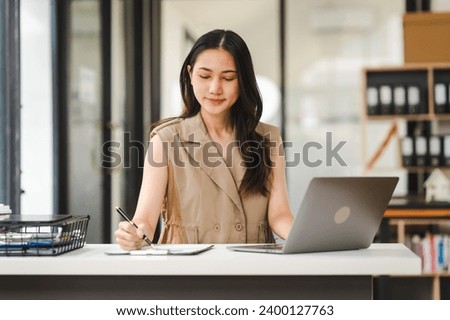 A focused Asian freelance writer takes notes at her desk with a laptop in a bright, modern office setting, journalist, artist, portrait, Thai's people. Royalty-Free Stock Photo #2400127763