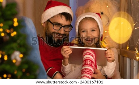 Happy father and little daughter in red santa hats use tablet and laugh sitting by Christmas tree. Family leisure time during holidays New Year and Christmas