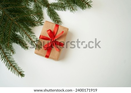 The gift on a white background is wrapped in craft paper with a red ribbon. Christmas tree branches are nearby. The concept of Christmas gifts