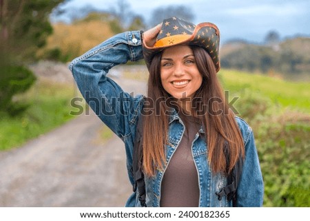 Smiling radiant woman with cowboy hat walking on a rural path while travelling alone