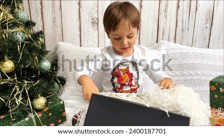 The boy throws the paper filler looking for a gift in a gift box. A cute little boy sitting on the bed near the Christmas tree open christmas present. Christmas spirit.