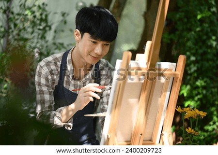 Happy Asian male sitting in sunny garden and painting picture with watercolor