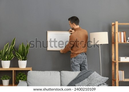 Handsome man hanging photo frame on dark wall at home, back view