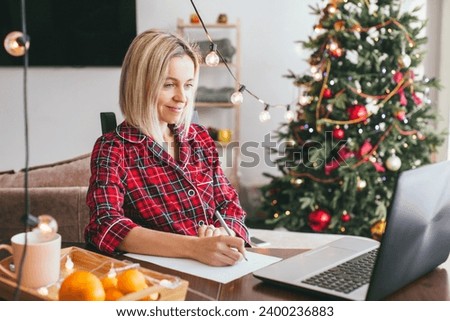 Middle aged woman works at home remotely with laptop on the background of Christmas tree. Work on holidays. Atmosphere and work at home