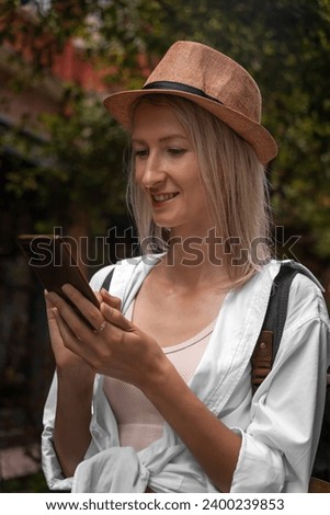 Beautiful young woman is walking around the city in a hat, a smiling stylish girl is holding a smartphone in her hands. Blonde tourist types a message and browses online pages on her mobile phone