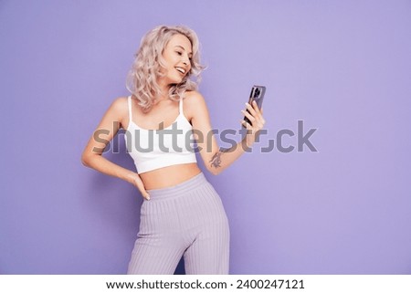 Young beautiful smiling female in trendy summer white top and pants clothes. Carefree blond woman posing near violet wall in studio. Positive model holds smartphone, uses phone apps, looks at screen 