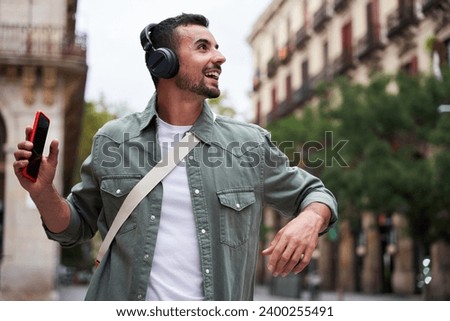 Young caucasian man having fun while listening to music on his headphone in the street on the city. Cheerful guy dancing outdoor looking to the side while holding his mobile in his hand. Royalty-Free Stock Photo #2400255491