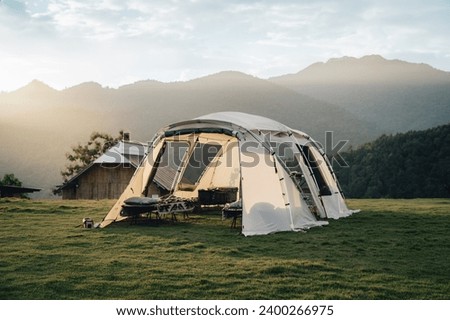 A big white camping tent with camping gears in a green grass with mountains and blue sky with white clouds in the background. Taken during golden hour Royalty-Free Stock Photo #2400266975