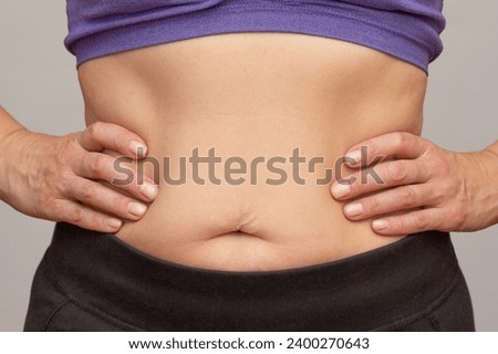 Cropped woman body with hands on waist showing belly sagging skin after diet and stretch marks after pregnancy over gray background Royalty-Free Stock Photo #2400270643