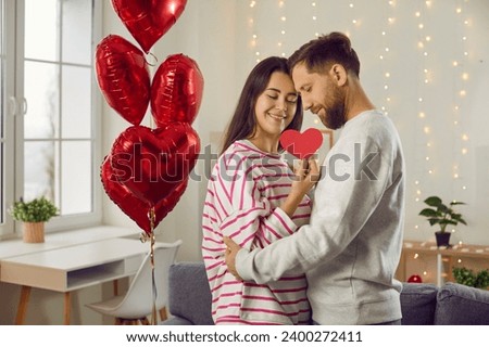 Portrait of a young brunette man and woman holding heart in her hands,hugging with closed eyes indoor with red foil ballons. Happy couple in love. Care, tenderness and Valentines day concept.