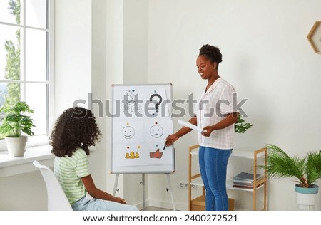 School counselor or psychologist working with a little girl, standing by a white board with pictures, pointing at a thumbs up symbol and asking the child questions. Children's psychology concept