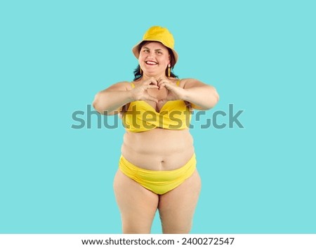 Portrait of a funny young happy fat woman wearing yellow beach bikini and hat doing heart symbol shape with hands isolated on studio blue background. Vacation trip and summer travel concept.
