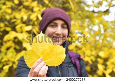 A woman in a burgundy hat and backpack holds out autumn leaves and smiles.  Beauty is in nature. Space for text. Focus on the foreground. Royalty-Free Stock Photo #2400277299
