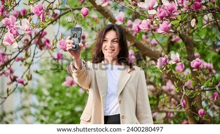 Smiling woman taking selfie with smartphone among spring blossoms with soft focus on the background.