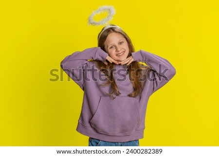 Portrait of smiling shy angelic young preteen child girl kid with angel halo nimb over head flirting, looking at camera, positive emotions, celebrating holiday. Children isolated on yellow background Royalty-Free Stock Photo #2400282389