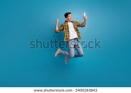 Full size body photo of funny man in air jumping with smartphone makes selfie shows his emotions isolated on blue color background