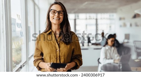 Business woman smiling at the camera while holding a tablet pc. Mature business woman standing in an office with her colleagues working in the background.