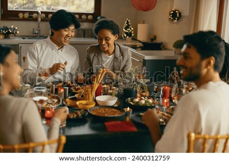 Group of friends sitting around festive dinner table at home, eating and looking at mobile phone