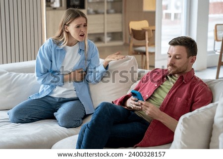 Upset indignant woman and ignoring man looking at phone screen while sitting on sofa at home. Dissatisfied unhappy wife demands attention from smartphone addicted husband. Stressed family conversation