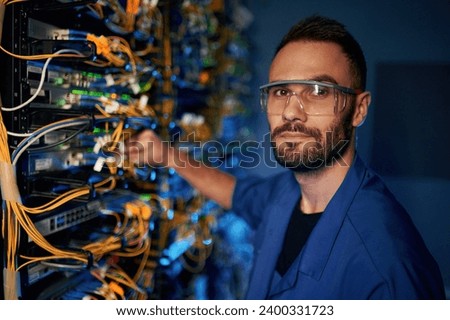 Looking to the camera. Young man is working with internet equipment and wires in server room. Royalty-Free Stock Photo #2400331723